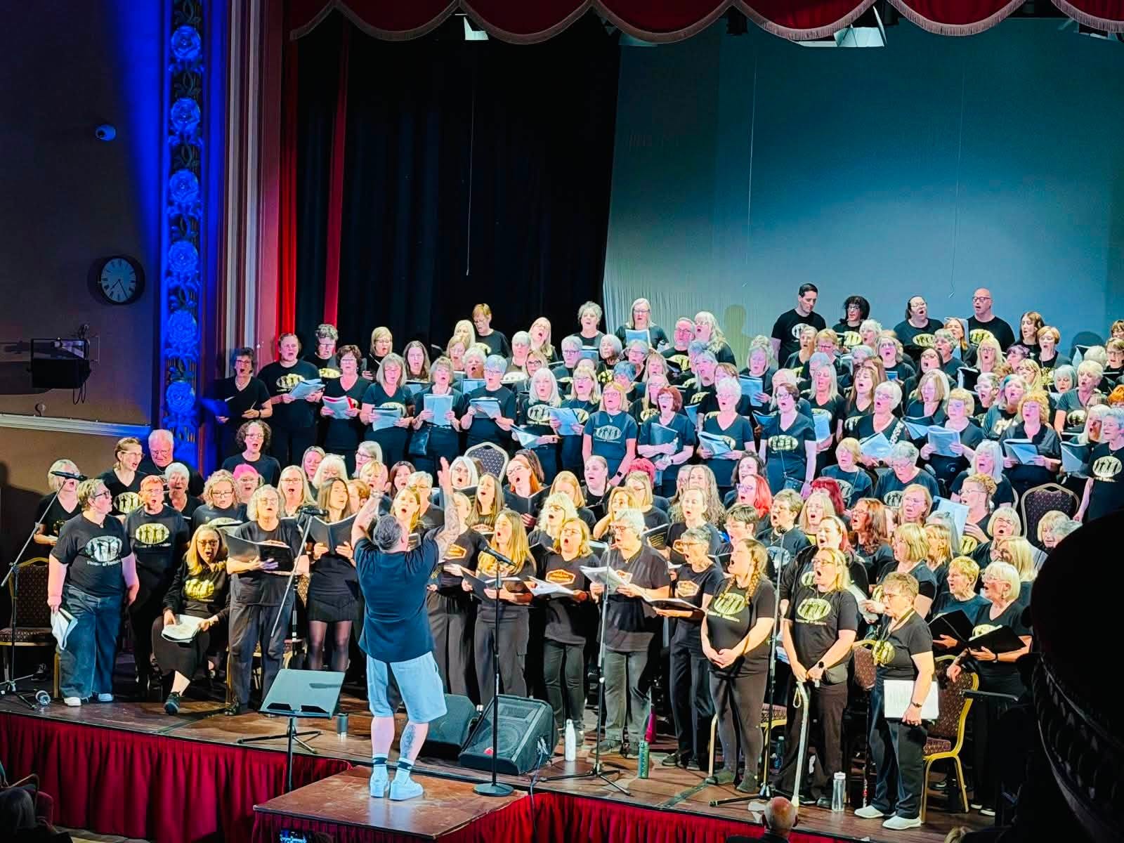 The full Voices of Yorkshire choir singing on a theatre stage with Nick conducting