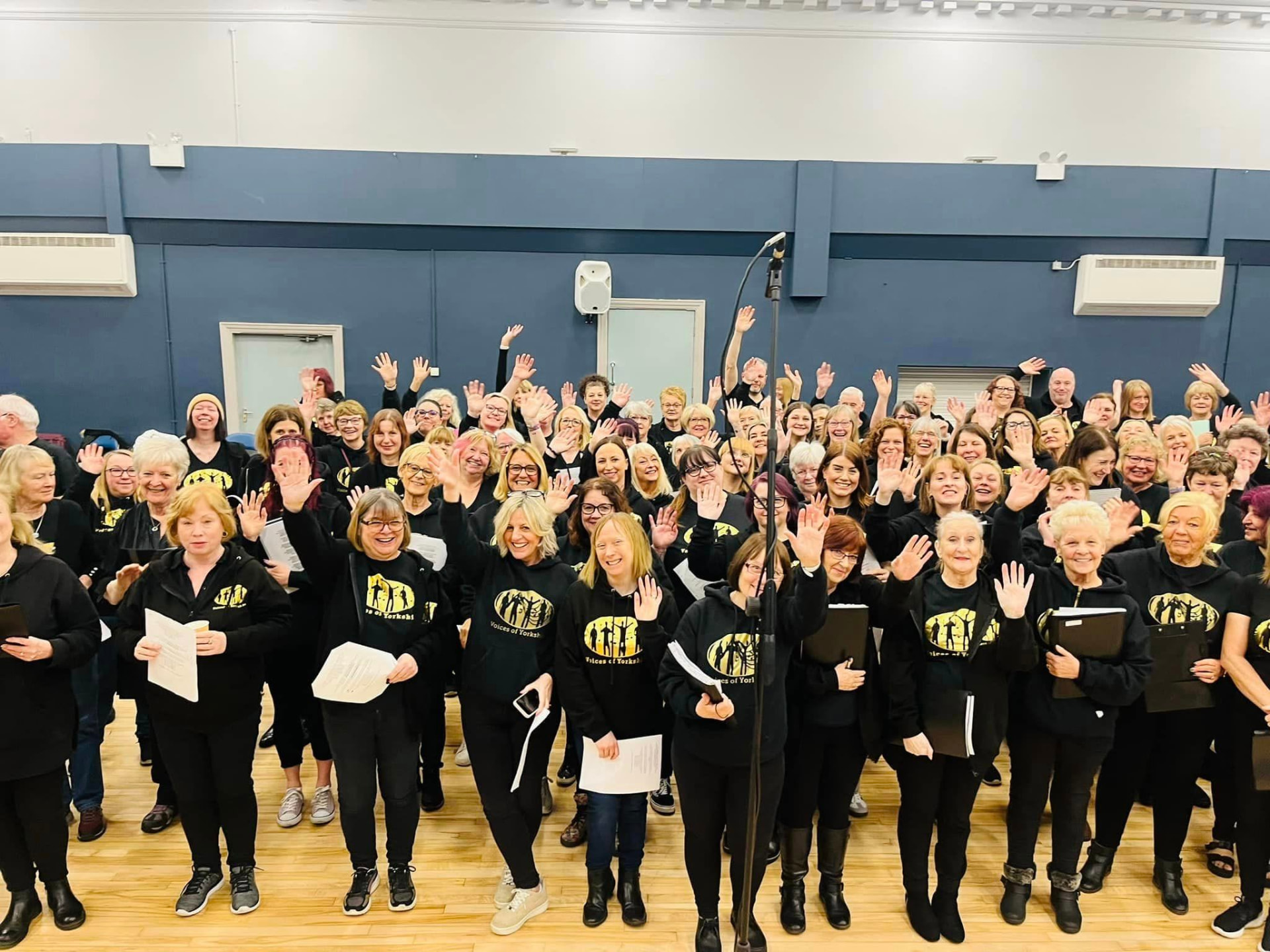 VOY members waving and smiling in their black and gold hoodies at a rehearsal